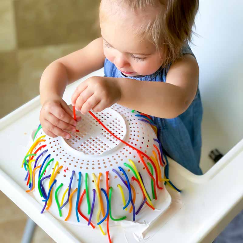 Rainbow spaghetti sensory play baby girl pulls spaghetti from colander for fine motor development