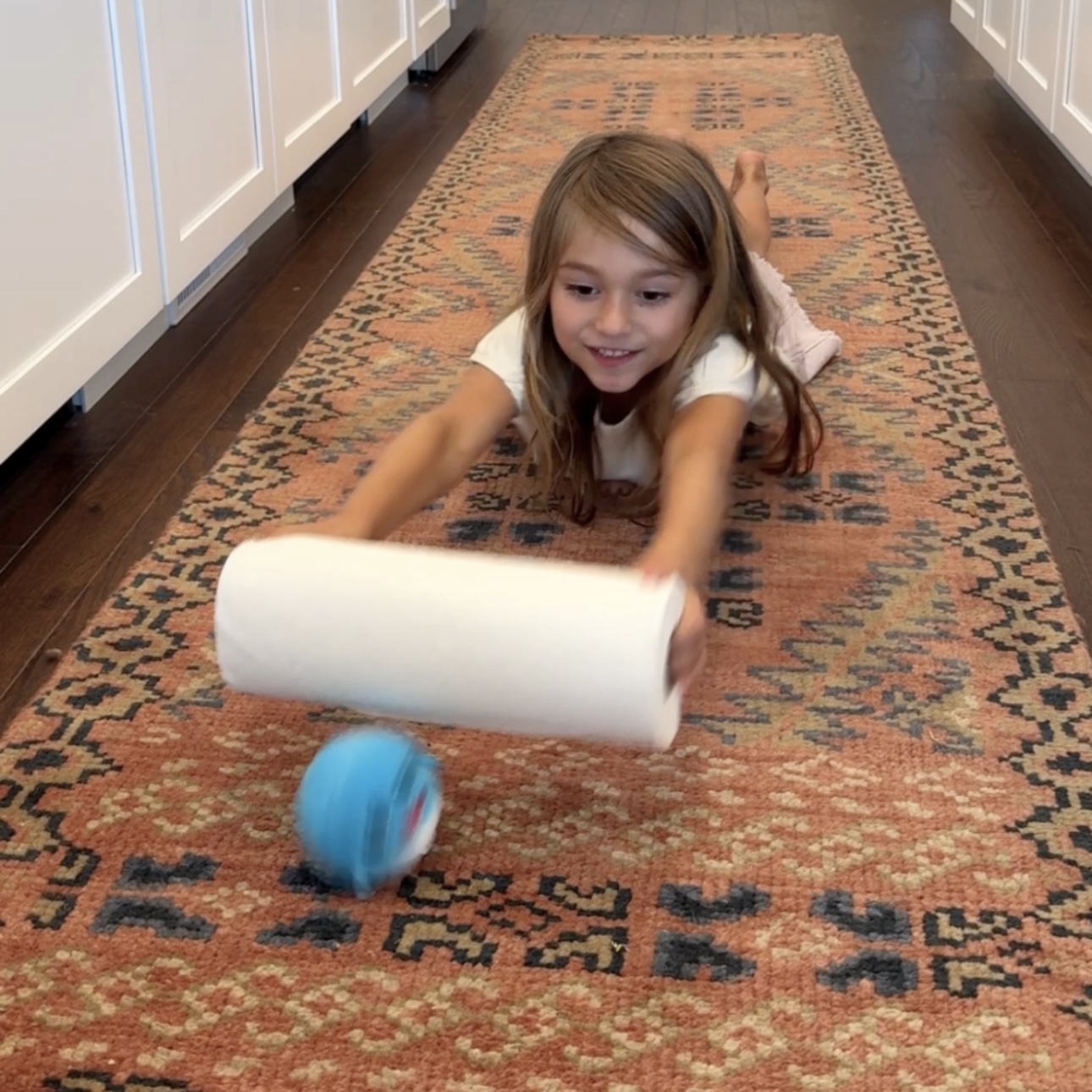 Child lying on stomach using a paper towel roll to push a plush ball forward