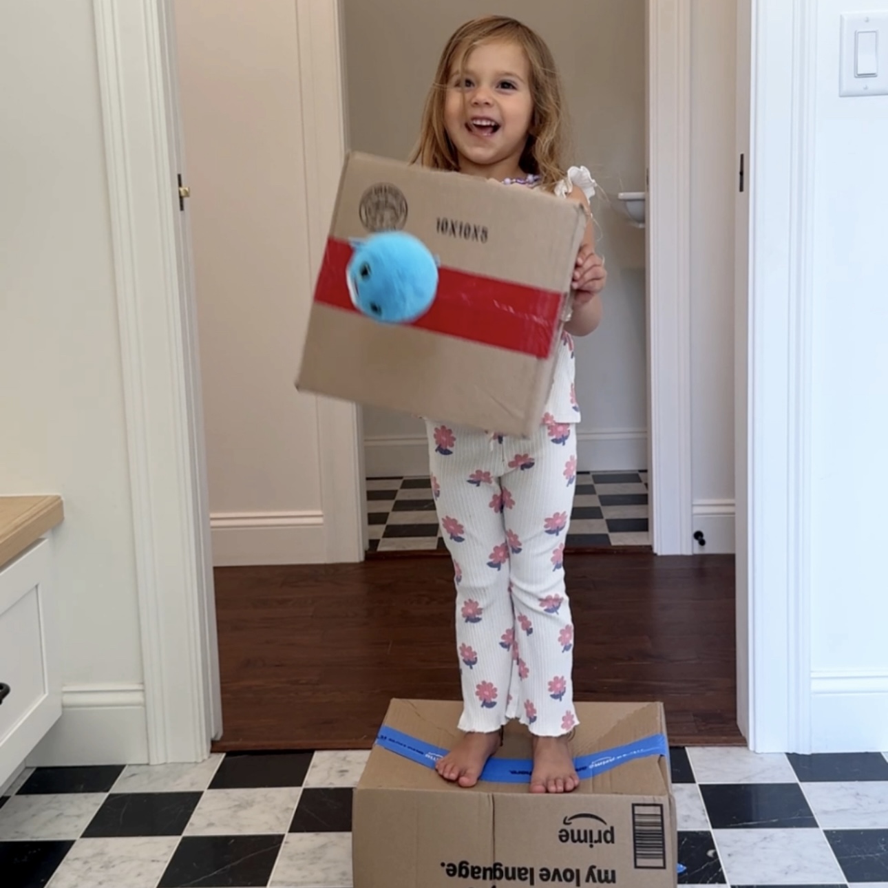 Child standing on a cardboard box balancing while holding a second box to block incoming plush balls