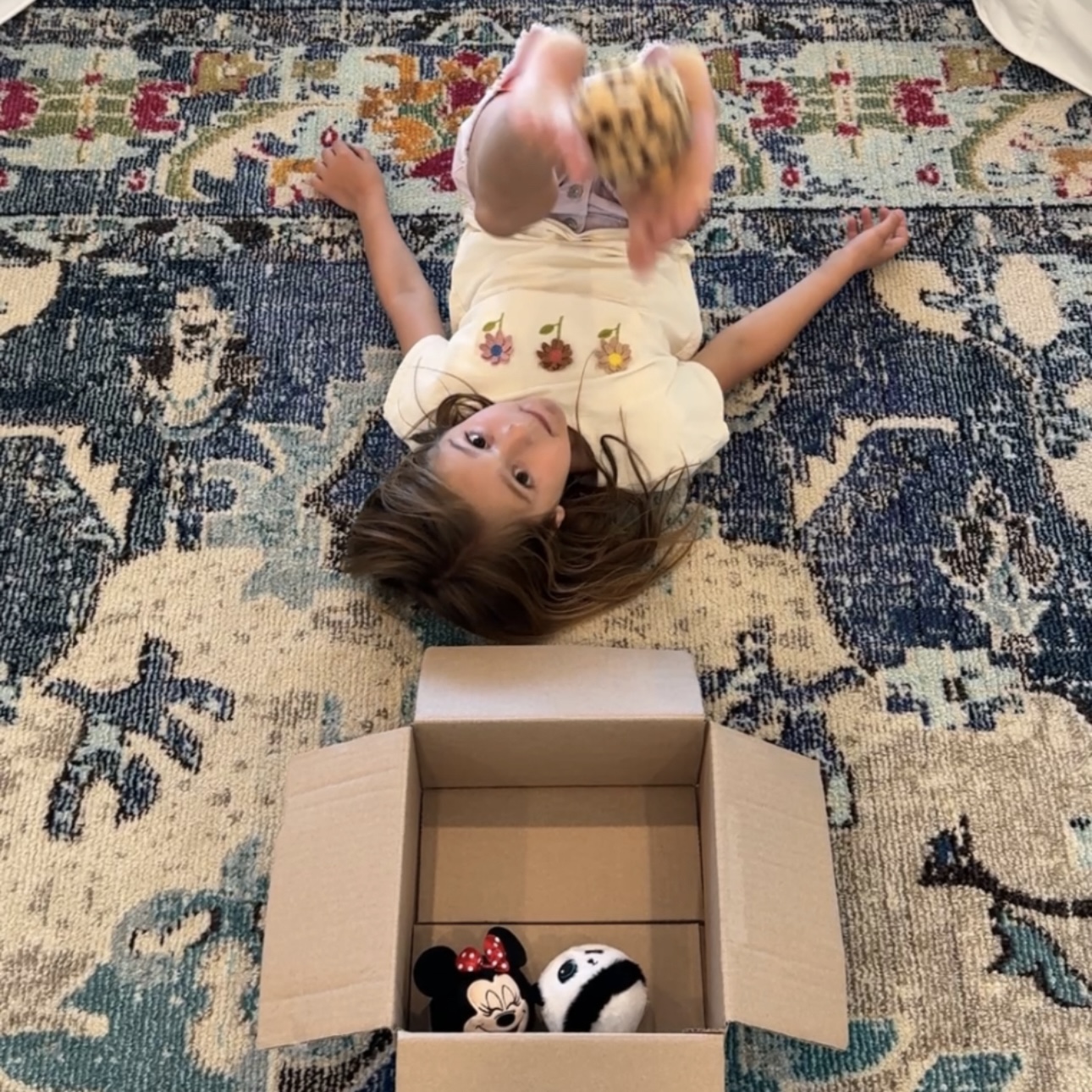 Child lying on back lifting a plush ball with feet and dropping it into a cardboard box