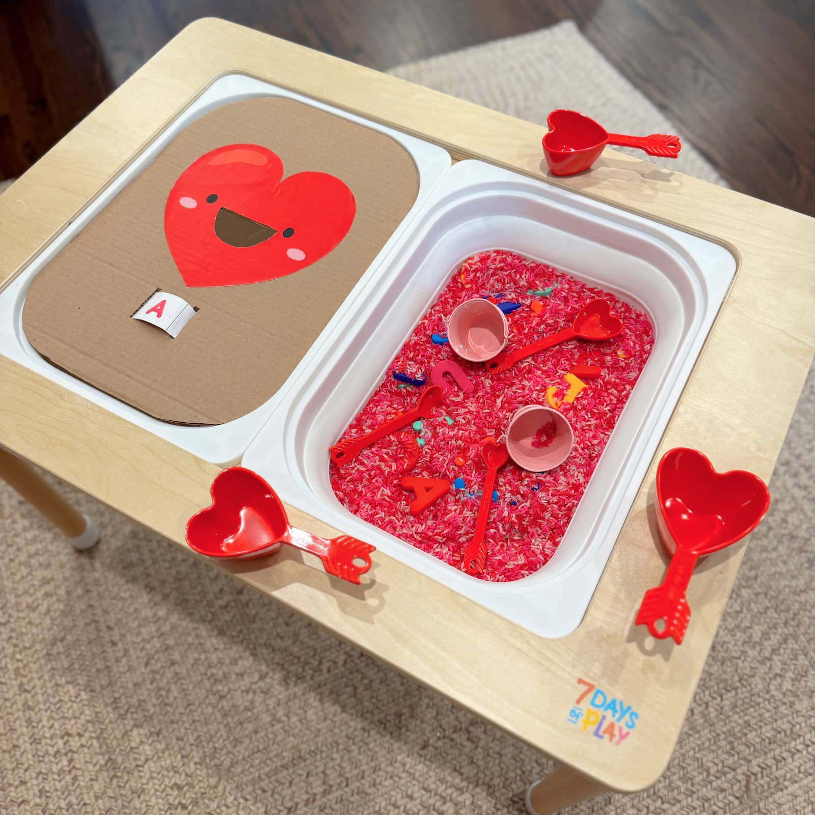 Close-up of a cardboard heart with a letter slot surrounded by pink Valentine’s sensory rice at a kids sensory table.
