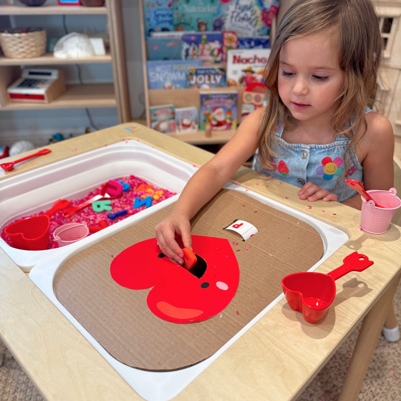 Child Feeding Letters to the Heart Child placing a plastic letter into a cardboard heart during a Valentine’s sensory rice activity at a sensory table.