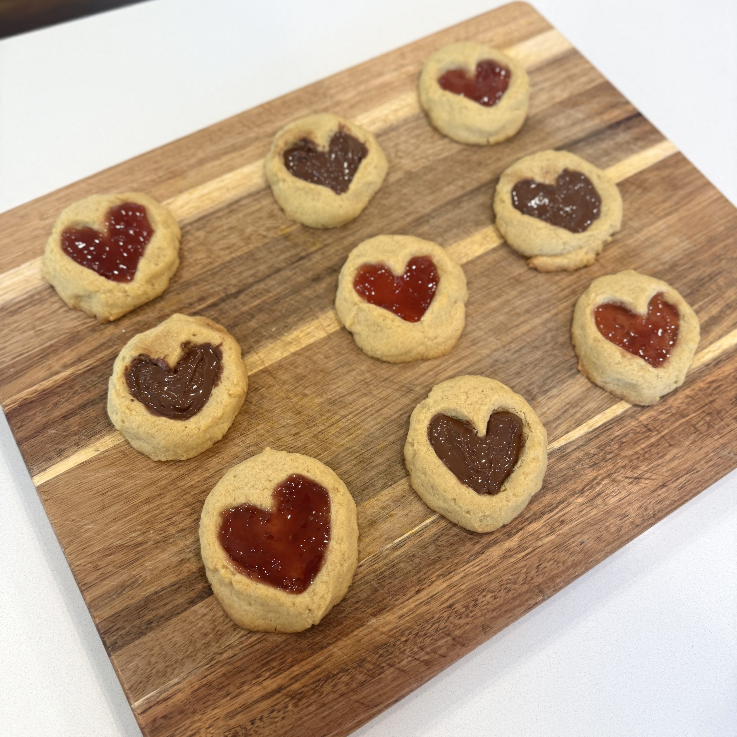 Thumbprint heart cookies on a wooden cutting board, filled with raspberry jam and Nutella.