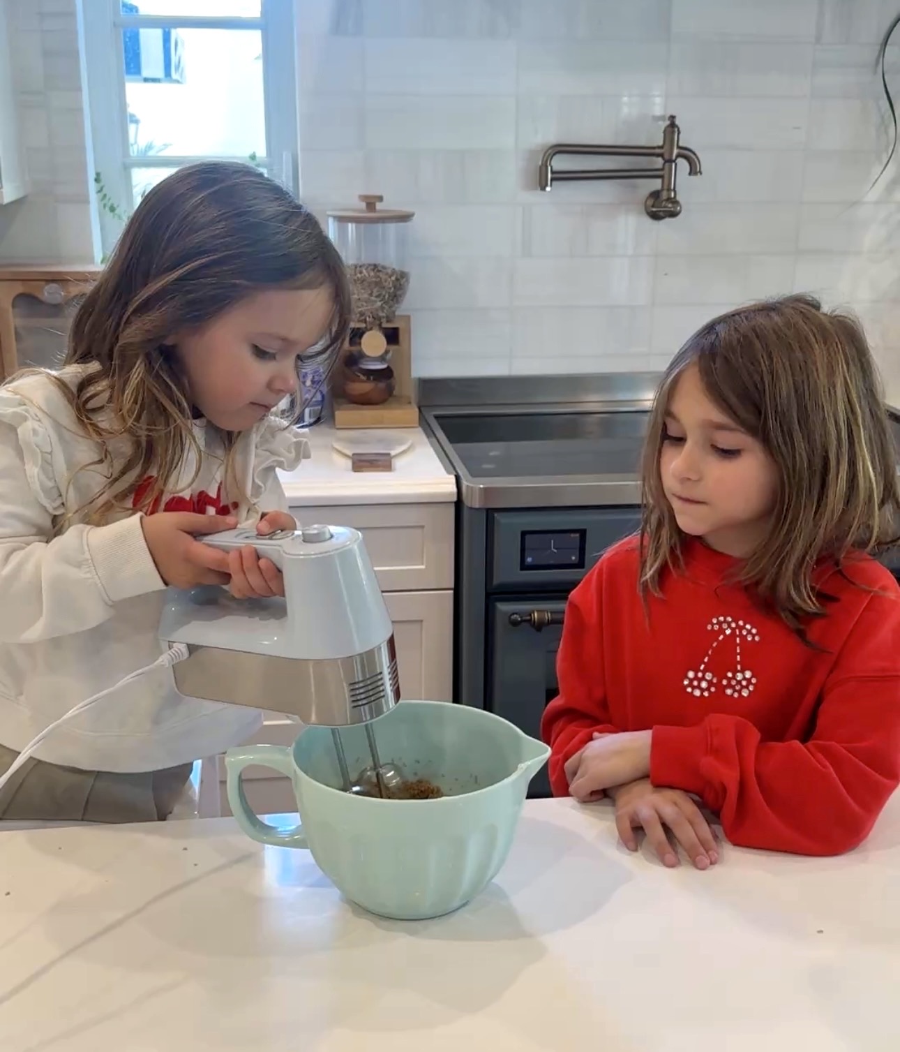 Two young children baking in a kitchen, one using a hand mixer to blend cookie dough in a mixing bowl while the other watches.