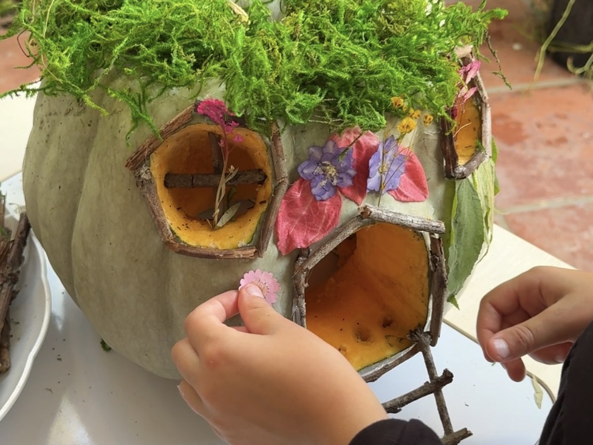 A child pressing a small pink flower onto a fairy house pumpkin decorated with twig window frames