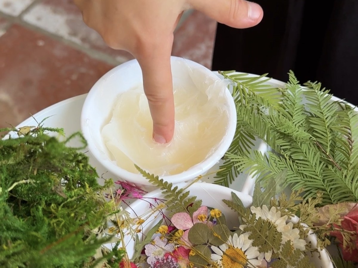 A child dipping a finger into a bowl of petroleum jelly beside a tray of pressed flowers and moss
