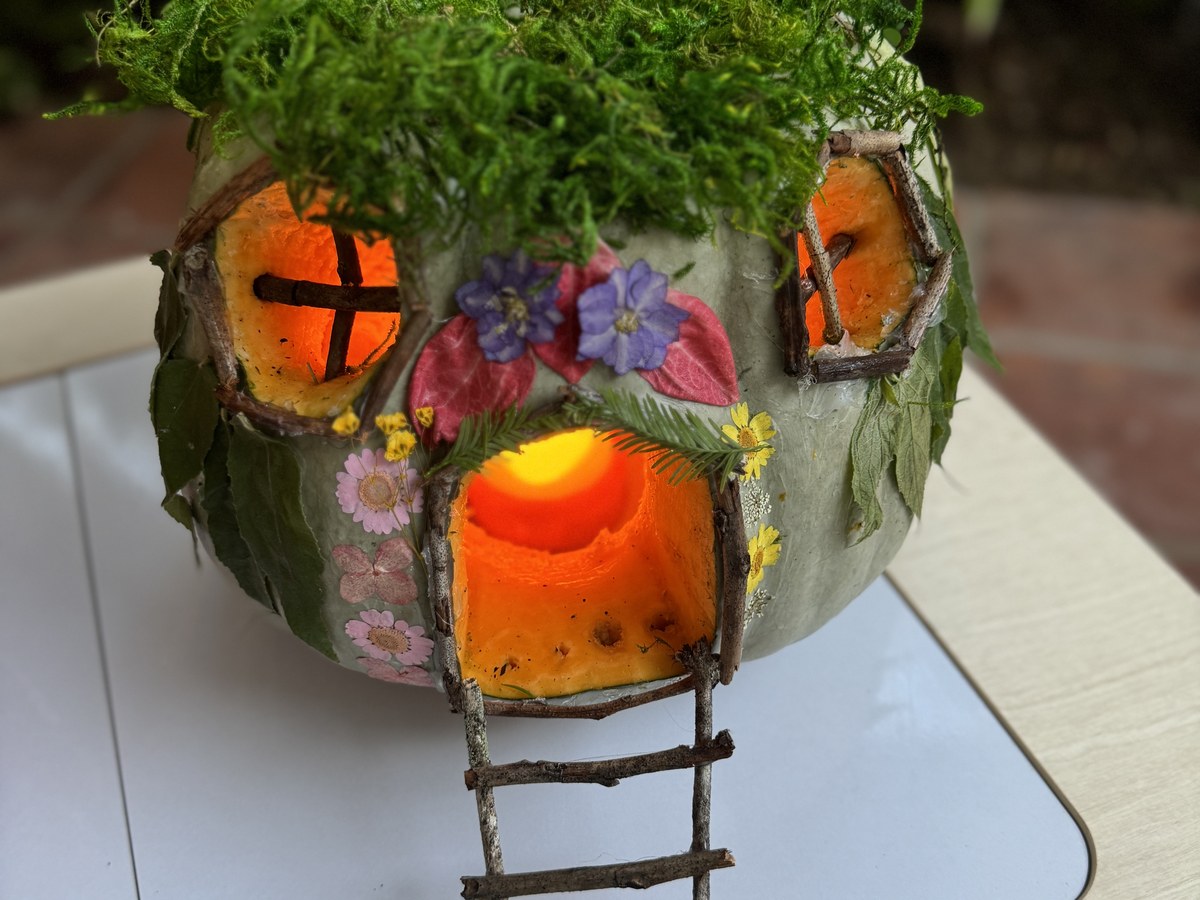 Close up of a fairy house pumpkin with a moss roof, twig window frames and a stick ladder up to the door