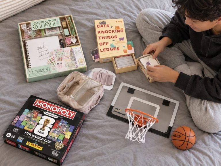 A teenager sitting on a bed opening a card game, with a DIY journal set, a travel makeup bag, a mini basketball hoop and a board game laid out on the duvet